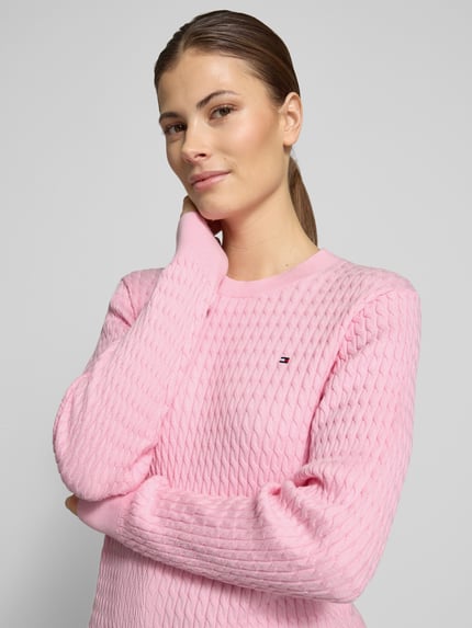 Vrouwenmodel draagt Slim Fit Pullover aus reiner Baumwolle van Tommy Hilfiger in effen roze met kabelpatroon, lange mouw, ronde hals, logodetail, gebreide look, puur katoen. Nahaufnahme bovenlichaam.