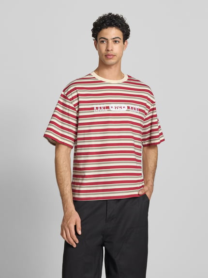 Männermodel trägt Relaxed Fit T-Shirt mit Logo-Stitching Modell 'Heritage' von KARL KANI, gestreift in Rot und Creme, Reine Baumwolle, Rundhalsausschnitt, Rückenlänge bei Größe M 64 cm, Herren T-Shirt, Ganzkörperansicht von vorne.