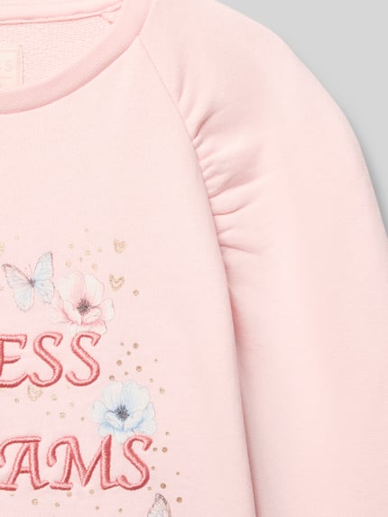 Dziewczynka nosi bluzę Sweatshirt mit Label-Detail od Guess z bawełny, krój regular fit, prążkowane wykończenia, okrągły dekolt, detal z logo, jasnoróżowa, kwiatowy wzór, miękka faktura, detal haftu, nahaufnahme górnej części.