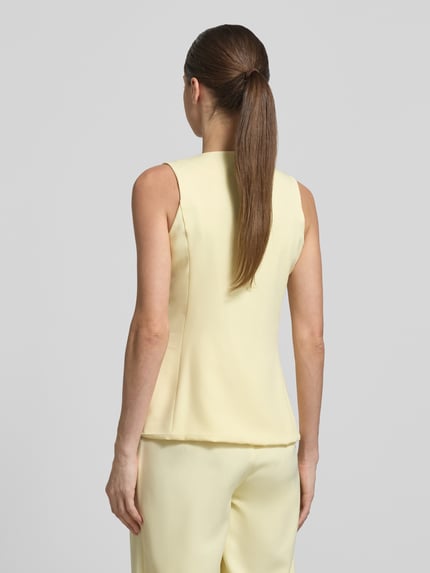 Frauenmodel trägt Weste mit Rundhalsausschnitt und Paspeltaschen von Little Mistress, Material-Mix mit Stretch-Anteil, Slim-Fit, Rückenlänge bei Größe 36: 61 cm, sanftes Gelb mit glatter Textur, Ganzkörperansicht Rückansicht.