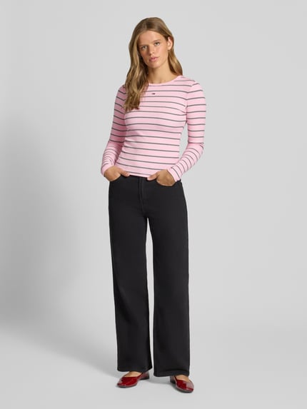 Frauenmodel trägt Slim Fit Langarmshirt aus Baumwoll-Mix von Tommy Jeans, rosa mit schwarzen Streifen, kombiniert mit schwarzer Hose, Ganzkörperansicht von vorne, Damen Langarmshirt von Tommy Jeans.