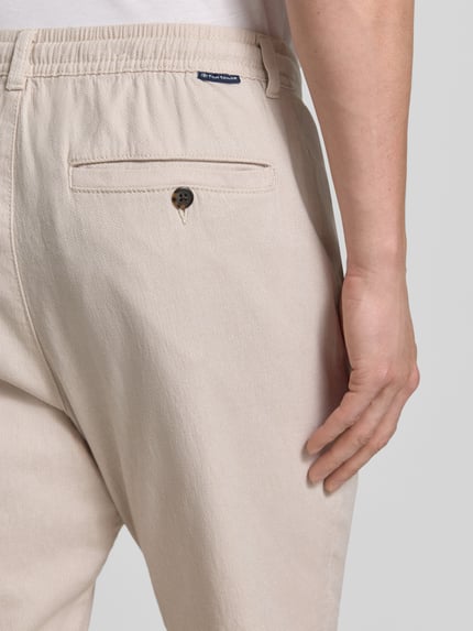 Mężczyzna nosi Regular Fit Chino aus Baumwoll-Mix von Tom Tailor, beżowy melanż, detal tylnej kieszeni, bawełniany miks, krój regular fit, label patch, szlufki na pasek, zapięcie na guzik i kryty zamek, tylne oraz boczne kieszenie, widok z tyłu, zbliżenie na górę.