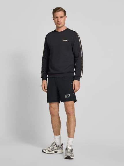 Männermodel trägt schwarze Shorts mit Logo und elastischem Bund von EA7 Emporio Armani, Material-Mix, Regular Fit, Ganzkörperansicht von vorne