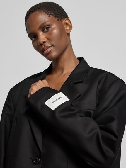 Frauenmodel trägt Regular Fit Longblazer mit Label-Detail von black palms, unifarben, Reverskragen, 1-Knopf, lange Ärmel, Nahaufnahme Oberkörper.