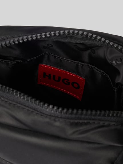 Produktfoto, czarny plecak z aplikacją z metką model 'AYDEN_MONOSTRAP' od HUGO, jednolity kolor, zamek błyskawiczny, główna przegroda, przegródka, przegródka zapinana na zamek błyskawiczny, możliwość noszenia na jedno ramię