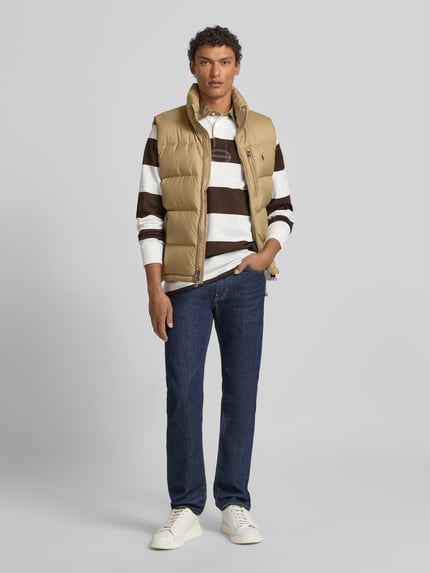 Männermodel trägt Sweatshirt mit Label-Stitching von Polo Ralph Lauren aus reiner Baumwolle, Regular-Fit, Polokragen, Knopfleiste, gestreiftes Muster in Braun und Weiß, gerippte Ärmelabschlüsse, kombiniert mit Weste und Jeans, Ganzkörperansicht von vorne.