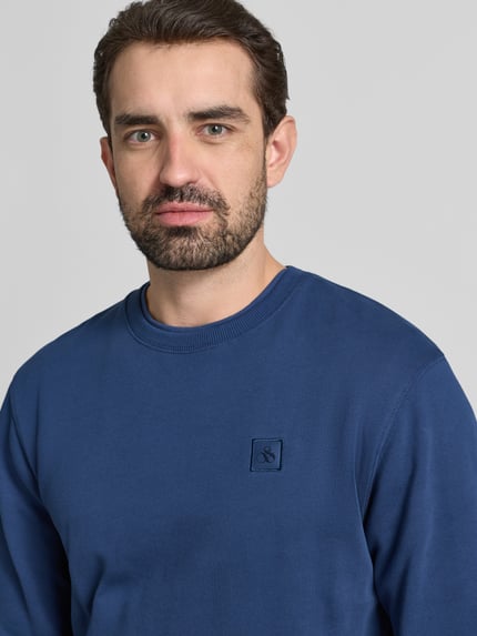 Männermodel trägt Sweatshirt mit Label-Detail von Scotch & Soda in Blau, reine Baumwolle, lockerer Schnitt, Nahaufnahme Oberkörper.