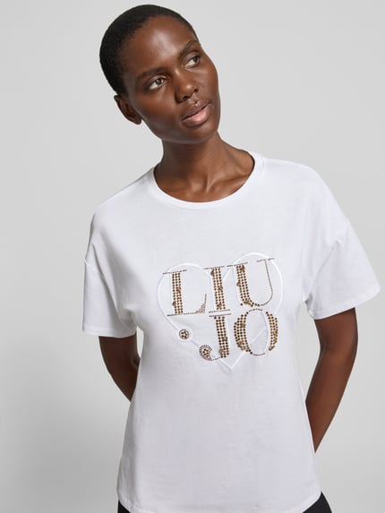 Kobieta nosi Regular Fit T-Shirt mit Strasssteinbesatz von Liu Jo White z bawełny, okrągły dekolt, detal z logo, długość tyłu 60 cm, zbliżenie na górną część ciała.