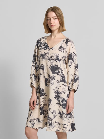 Frauenmodel trägt Fließendes Tunikakleid aus reinem Leinen von Marc O'Polo, Optik:Allover-Muster, Passform fließend, Midi-Länge, V-Ausschnitt, floral, Kurzarm, Ärmelabschluss elastisch, Materialverarbeitung Flammgarn, Material reines Leinen, cremefarben mit dunklem Blumen-Muster, strukturierte Oberfläche, Ganzkörperansicht von vorne.