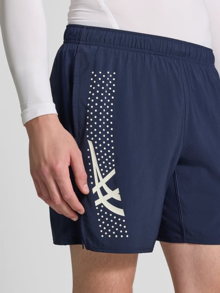 Männermodel trägt Regular Fit Shorts mit integriertem Slip Modell 'ICON' von ASICS TIGER in Dunkelblau mit Logo-Print, Nahaufnahme Oberschenkel, elastischer Bund, Kontext: integrierter Slip, Bundweite 37 cm, Innenbeinlänge 18 cm.