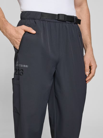 Mężczyznamodel nosi spodnie typu track pants model 'HALO MOVE' od HALO, mieszanka materiałów z dodatkiem streczu, krój regular fit, nadruk z logo, kieszenie zapinane na zamek błyskawiczny, długość wewnętrzna nogawki w rozmiarze M: 78 cm, ciemny kolor, lekko połyskująca struktura, detaliczny widok na górną część nogawki z przodu