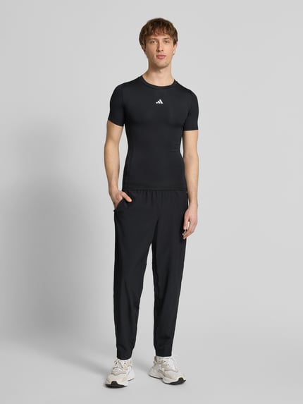 Mężczyzna-model nosi T-Shirt mit Label-Print od adidas Training, czarny materiał z elastycznym dodatkiem, krój slim fit, góra z przodu, zdjęcie całopostaciowe z przodu.