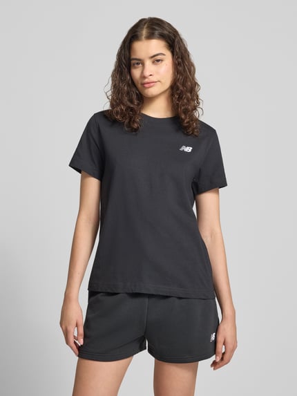 Frauenmodel trägt Regular Fit T-Shirt mit Logo-Stitching von New Balance, Damen T-Shirt aus Reine Baumwolle, Rundhalsausschnitt, Rückenlänge 60 cm, schwarze Farbe, glatte Struktur, Ganzkörperansicht von vorne.