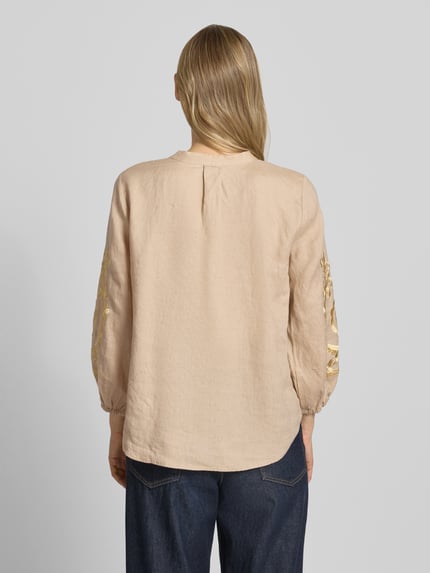 Frauenmodel trägt Leinenbluse mit Motiv-Stitching und Quasten-Details von Emily Van den Bergh in Beige, elastische Ärmelabschlüsse, Loose-Fit, reine Leinenqualität, Rückenlänge 64 cm, Ärmellänge 50 cm, Ganzkörperansicht Rückansicht.