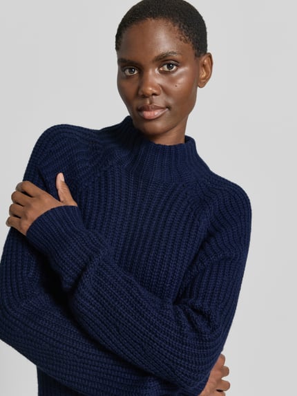Frauenmodel trägt dunkelblauen Strickpullover aus Kaschmir-Baumwoll-Mix Modell 'LONA' von 0039 Italy, grobe Struktur, Nahaufnahme Oberkörper, Damen Regular Fit, Stehkragen, Rückenlänge 60 cm, Ärmellänge 78 cm.