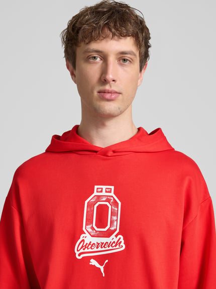 Męski model nosi Relaxed Fit Hoodie mit Känguru-Tasche od PUMA PERFORMANCE, czysta bawełna, detal z logo, kolor czerwony, zbliżenie na górną część ciała.