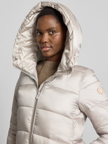 Frauenmodel trägt Steppmantel mit Kapuze Modell 'LYSA' von Save The Duck in Beige, glatte gesteppt-glänzende Oberfläche, Label-Patch, Nahaufnahme Oberkörper. Kontext: Gerader Schnitt, Zweiwege-Reißverschluss, seitliche Eingrifftaschen, Rückenlänge 91 cm, Schulterbreite 39 cm, Brustweite 40 cm, Ärmellänge 66 cm.