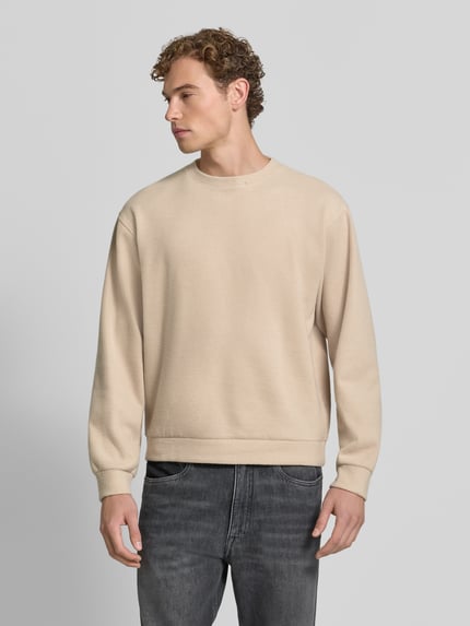 Męski model nosi beżowy sweter z okrągłym dekoltem Model 'KARIO' od DRYKORN, mieszanka bawełny, luźny krój, elastyczne wykończenia, długość tyłu w rozmiarze M: 70 cm, długość rękawa w rozmiarze M: 64 cm, widok całej sylwetki z przodu.