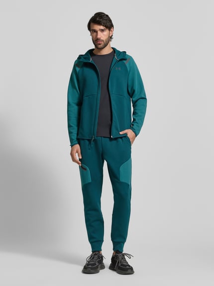 Męski model nosi Regular Fit Spodnie dresowe z logo-Print model 'Unstoppable' od Under Armour, elastyczna mieszanka materiałów z dodatkiem bawełny, kieszeń na nogawce z zamkiem błyskawicznym, elastyczny pas i zakończenia nogawek, długość wewnętrzna nogawki w rozmiarze M: 73 cm, kolor morski, gładka faktura, krój regular fit, całkowity widok z przodu.