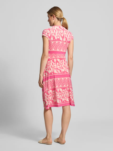 Frauenmodel trägt knielanges Kleid mit V-Ausschnitt Modell 'Carrara' von ZABAIONE, elastischer Material-Mix, taillierte Passform, kurzärmeliges Design in Pink mit weißen Mustern, Raffungen, Rückenlänge 102 cm, Brustweite 46 cm, Ganzkörperansicht Rückansicht.