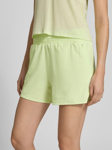 Modelka kobieca ma na sobie szorty Regular Fit Shorts z elastycznym pasem model 'Kout' od Salomon, szorty damskie z wbudowanymi figami, krój regular fit, szerokość w rozmiarze S: 36 cm, długość wewnętrzna nogawki w rozmiarze S: 10 cm, jasny kolor, widok całej sylwetki z przodu.