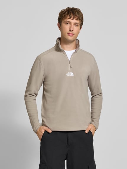 Männermodel trägt Fleecepullover mit Label-Stitching von The North Face in Beige, Regular Fit, Stehkragen, Reißverschluss, Ganzkörperansicht von vorne.