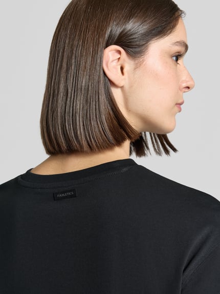 Frauenmodel trägt Relaxed Fit T-Shirt aus Baumwoll-Mix von Fabletics, schwarzer Stoff, glatte Textur, Nahaufnahme Oberkörper, Rückenansicht.