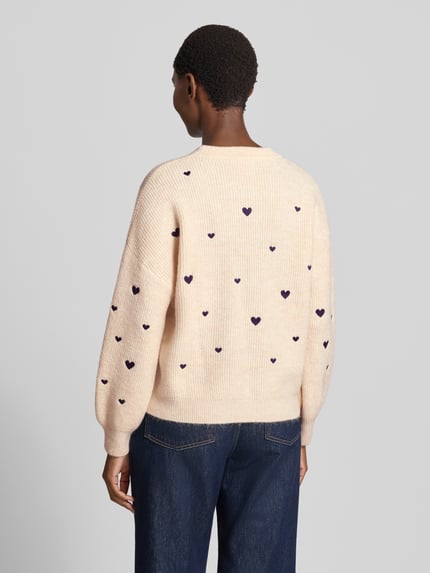 Frauenmodel trägt Strickpullover mit Woll-Anteil von Apricot in Beige mit violetten Herz-Stitchings, Ripp-Optik, Ballonärmel, Rückenansicht.