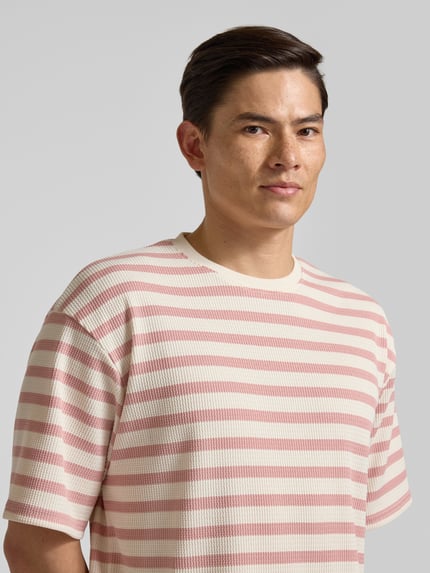 Männermodel trägt Oversized T-Shirt mit Rundhalsausschnitt Modell 'Gael' von Redefined Rebel, Elastischer-Material-Mix, Oversized-Fit, rosa-weiß gestreift, Nahaufnahme Oberkörper.