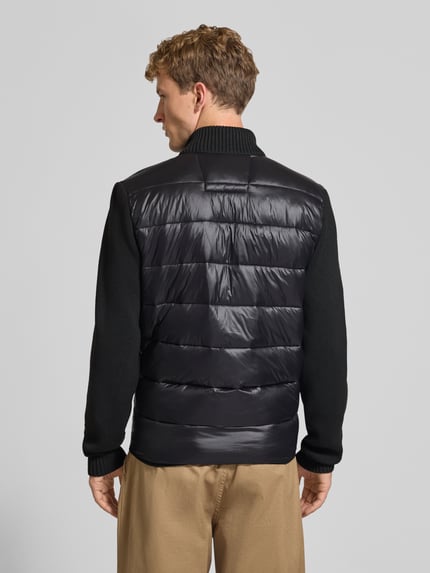 Mannenmodel draagt Leichtsteppjacke met gebreide mouwen en opstaande kraag van REVIEW, zwarte kleur, regular fit, labelstitching, ritssluiting, steekzakken, ruglengte maat M 70 cm, schouderbreedte 46 cm, borstwijdte 52 cm, mouwlengte 73 cm, Ganzkörperansicht Rückansicht.