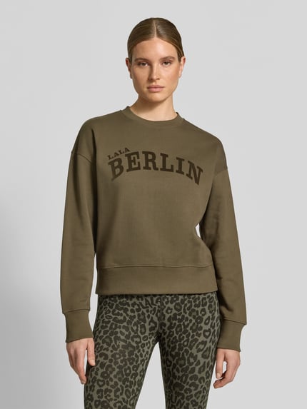 Frauenmodel trägt Sweatshirt mit Label-Print von Lala Berlin in Olivgrün, reine Baumwolle, lockerer Schnitt, Rundhalsausschnitt, Ganzkörperansicht von vorne.