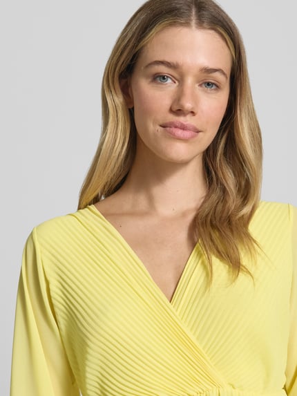 Frauenmodel trägt knielanges Kleid mit Plisseefalten Modell 'Im44sa' von ZABAIONE in Gelb, Wickel-Optik, Nahaufnahme Oberkörper.