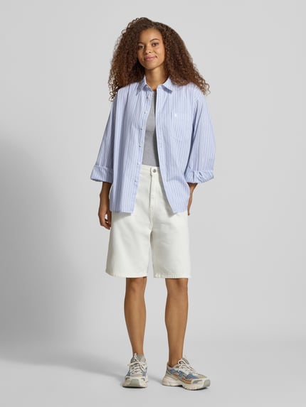 Vrouwenmodel draagt Relaxed Fit blouse met labelstitching Model 'Keene' van Carhartt Work In Progress, puur katoen, blousekraag, knoopsluiting, borstzak; kleuren lichtblauw met witte strepen, wit shirt, witte korte broek, lichte sneakers; context: dames blouse van Carhartt Work In Progress, puur katoen, relaxed fit, model 'Keene', overhemdkraag, knoopsluiting, borstzak, labelstitching, ruglengte bij maat S: 78 cm, mouwlengte bij maat S: 56 cm. Volledig lichaamsaanzicht van voren.