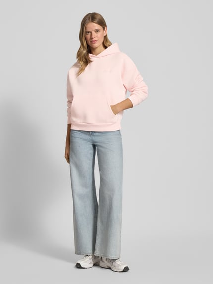 Frauenmodel trägt Oversized Hoodie mit Kapuze von Review in zartem Rosa, aus Baumwoll-Mix mit Label-Print und Känguru-Tasche, Ganzkörperansicht von vorne.