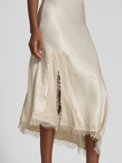 Frauenmodel trägt Maxikleid mit Spitzenbesatz von VERO MODA, unifarben, glänzend, Satin, Spitzendetails, asymmetrischer Saum, Nahaufnahme Unterkörper.
