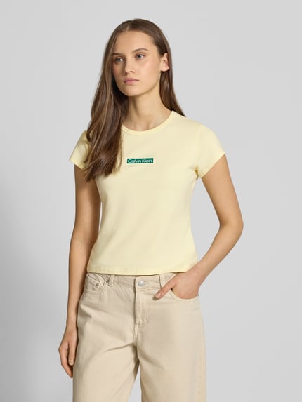 Modelka nosi T-shirt mit Rundhalsausschnitt und Label-Print von Calvin Klein Jeans, mieszanka bawełny i elastanu, krój slim fit, okrągły dekolt, nadruk z logo, długość tyłu w rozmiarze S: 42 cm, jasny kremowy kolor, widok całej sylwetki z przodu.