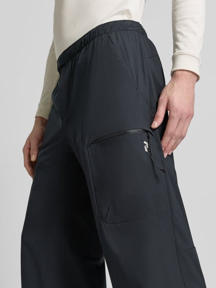Mężczyzna nosi Regular Fit Hose mit Reißverschlusstaschen od Peak Performance, czarny, elastyczny pas, krój regular fit, kieszenie zapinane na zamek błyskawiczny, Szerokość w rozmiarze M: 40 cm, Długość wewnętrzna nogawki: 76 cm, zbliżenie na dolną część ciała z przodu.