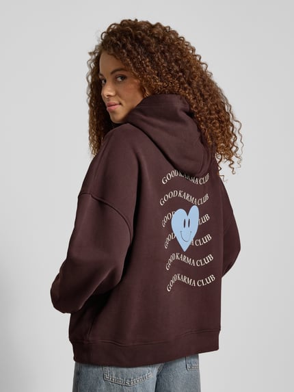 Frauenmodel trägt Hoodie mit rückseitigem Print Modell 'Good Karma Wave' von OH APRIL, Oversized, Baumwoll-Mix, dunkelbraun, Ganzkörperansicht Rückansicht.