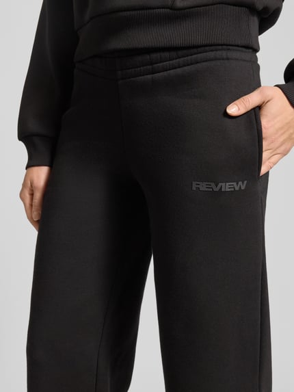 Vrouwenmodel draagt zwarte Sweatpants met elastische band van Review, losvallende pasvorm, labelprint, close-up heupen.