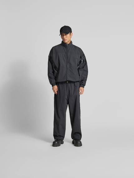 Männermodel trägt schwarze Trainingsjacke mit Stehkragen von VETEMENTS, Relaxed-Fit, Zweiwege-Reißverschluss, elastische Abschlüsse, Eingrifftaschen, Ganzkörperansicht von vorne. Rückenlänge 74 cm, Schulterbreite 50 cm, Ärmellänge 73 cm.