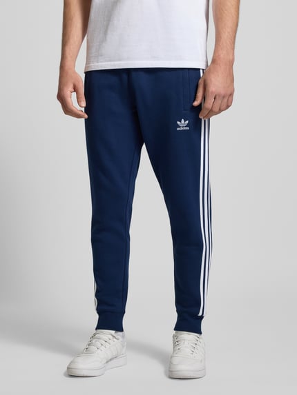 Mężczyzna nosi Spodnie dresowe z elastycznym pasem od adidas Originals, mieszanka bawełny, luźny krój, wpuszczane kieszenie, haftowane logo, długość wewnętrzna nogawki 38 cm, rozmiar M, Ganzkörperansicht von vorne.