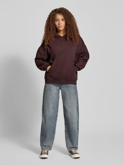 Frauenmodel trägt Hoodie mit rückseitigem Print Modell 'Good Karma Wave' von OH APRIL, dunkelbraun, Baumwoll-Mix, oversized, Känguru-Tasche, Ganzkörperansicht von vorne.