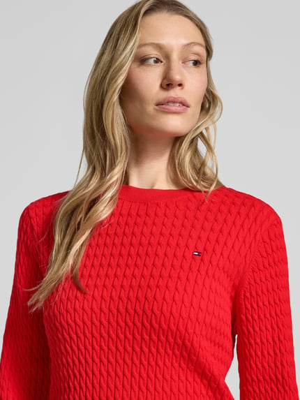 Frauenmodel trägt Slim Fit Pullover aus reiner Baumwolle von Tommy Hilfiger in Rot, Zopfmuster, Logo-Detail, Nahaufnahme Oberkörper.