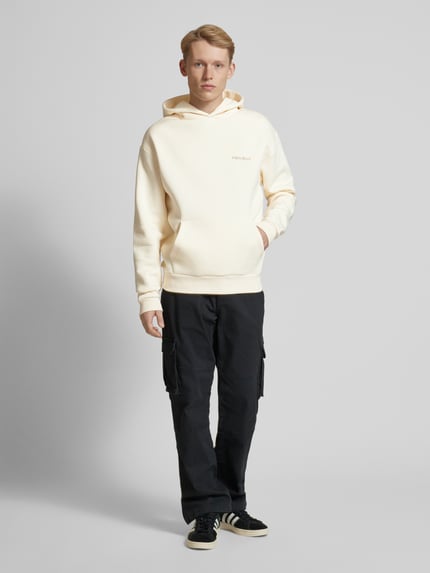 Männermodel trägt Hoodie mit Label-Print von REVIEW in Creme mit Känguru-Tasche, Baumwoll-Mix, schwarze Hose, Ganzkörperansicht von vorne. Rückenlänge 68 cm, Ärmellänge 67 cm, Kapuze und elastische Abschlüsse.