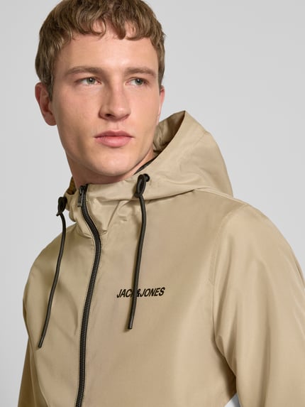 Männermodel trägt Jacke mit Kapuze und Reißverschluss Modell 'RUSH' von JACK & JONES in Beige, Logo, Regular-Fit, Nahaufnahme Oberkörper. Rückenlänge 68 cm, Schulterbreite 46 cm, Brustweite 55 cm, Ärmellänge 68 cm, mit Eingrifftaschen.