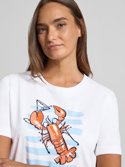 Frauenmodel trägt T-Shirt mit Motiv-Print und Pailletten von Marc Cain, Damen T-Shirt aus Reine Baumwolle in Weiß und Orange, Regular-Fit, Rundhalsausschnitt, Struktur akzentuiert durch Pailletten, Rückenlänge bei Größe 36: 56 cm, Nahaufnahme Oberkörper.