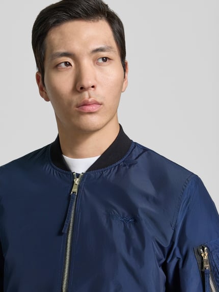 Männermodel trägt Bomberjacke mit Label-Patch und Eingrifftaschen von Original Bombers in Dunkelblau, Nahaufnahme Oberkörper. Gerader-Schnitt, Reißverschluss, Reißverschlusstasche am Ärmel, Druckknopfverschluss, Gerippte-Abschlüsse, Rückenlänge 67 cm, Schulterbreite 43 cm, Brustweite 53 cm, Ärmellänge 68 cm.