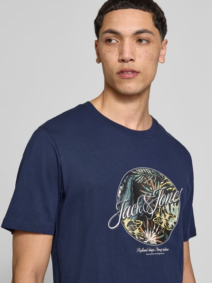 Męski model nosi t-shirt z nadrukiem Jack & Jones T-Shirt mit Rundhalsausschnitt Modell 'HONOLULU' z bawełny, krój regular fit, okrągły dekolt, długość tyłu w rozmiarze m: 68 cm, z przodu na wysokości klatki piersiowej, zbliżenie na górną część tułowia.
