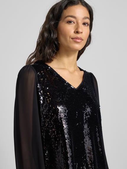 Kobiece model nosi Knielanges Kleid mit Paillettenbesatz von More & More, elastyczna mieszanka materiałów, taliowany krój, dekolt w serek, wycięcie w kształcie łezki z tyłu, zapięcie na guzik, półprzezroczyste bufiaste rękawy, długość do kolan, czarna połyskująca tekstura z cekinowym obszyciem, widoczna szerokość klatki piersiowej 50 cm, długość tyłu 92 cm, długość rękawa 60 cm, rozmiar 36, Nahaufnahme górnej części ciała.