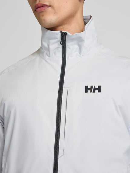 Mannenmodel draagt Bomberjacke mit Stehkragen Modell 'RACING' van Helly Hansen in lichtgrijs met zwarte ritssluiting, logoprint, recht model, opstaande kraag, steekzakken, ribboorden, detailafname bovenlichaam, Nahaufnahme Oberkörper.